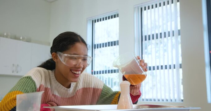 African-American schoolgirl leaning in, pouring orange beaker into flask for class, watching foam