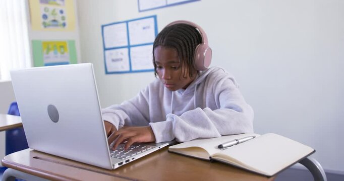 African American teen in gray hoodie, pink headset leaning typing on laptop in class checking notes