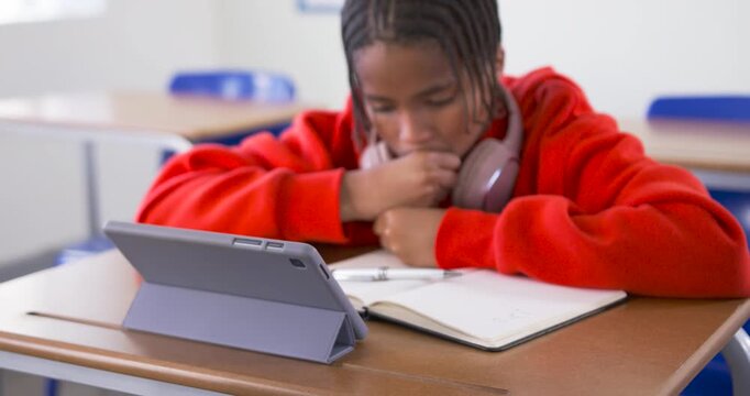 African teen glancing at tablet, picking pen at school desk and writing in notebook while studying
