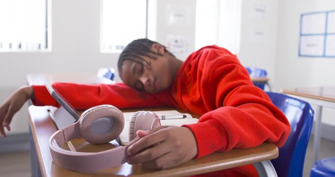 Teenage student in red hoodie resting head on school desk, adjusting headphones beside tablet
