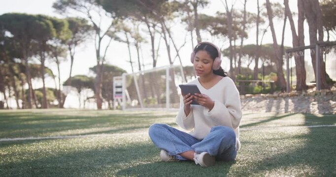 Female child sitting cross-legged starting looking down at tablet with pink headset tapping on turf