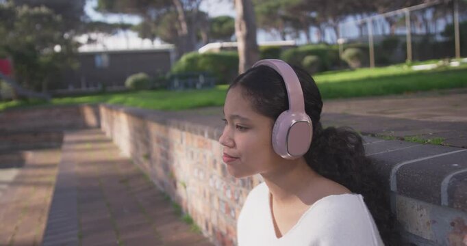 Teen sitting on brick wall at park, listening via pink headphones, mouthing to music, copy space