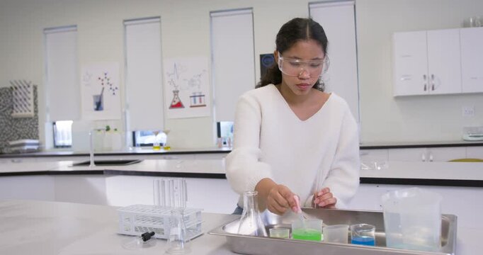 Schoolgirl with goggles reaching for tray drawing liquid with pipette mixing samples at lab bench