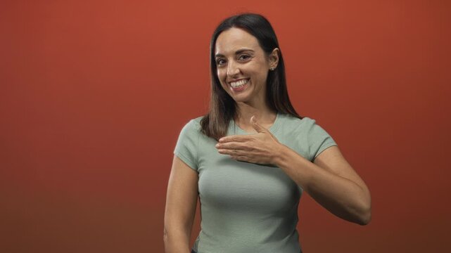 Woman touching hand to chest in studio red backdrop, smiling at camera, wearing casual t shirt and stud earring; warm gratitude.