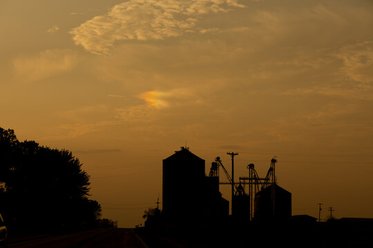 Darken silhouette of farm buildings and a road at dusk or dawn, with a sundog appearing overhead

