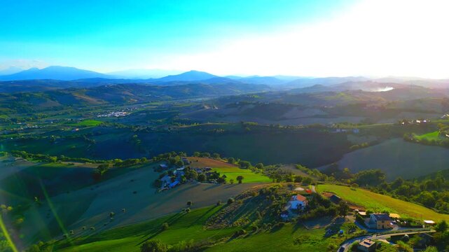 Splendid aerial truck left pan right shot from Monterubbiano, roads with cars winding over suave green-topped hills are imbued with the placid afternoon sunlight, over the lawns, fields, mountains
