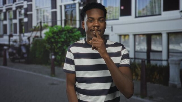 Young african american man wearing black and white striped shirt stands holding finger to lips for silence on busy street; quiet secrecy.