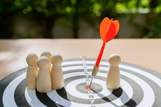 Dart in bullseye of dartboard and there are wooden figure standing around, representing a competition to reach a goal.