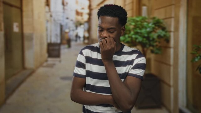 Man in striped shirt bites fingernails on narrow city street lined with stone walls under warm sunlight; anxiety uncertainty.