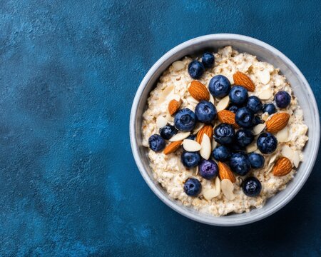 Bowl of oatmeal porridge topped with fresh blueberries sliced almonds on textured blue background representing healthy breakfast morning nutrition.