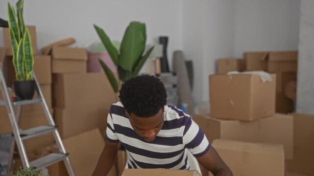 Man opens cardboard box in home office surrounded by moving boxes, potted plants, and a ladder; curiosity.