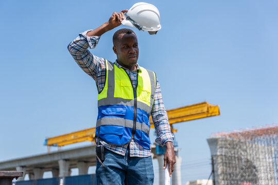African American engineer holding safety helmet and blueprints outdoors, Tired construction worker wiping sweat under the hot sun, Exhausted engineer taking a break at construction site in heat