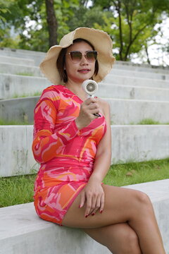Asian woman in a pink patterned dress and straw hat uses a small portable fan to stay cool outdoors