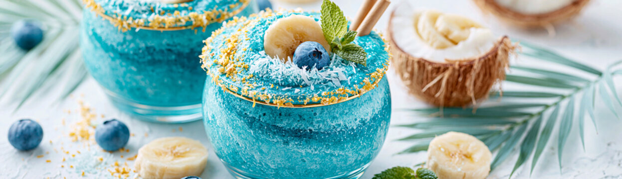 Blue smoothie bowls topped with blueberries, banana slices, coconut flakes, and mint leaves in glass jars, set on a light surface with coconut halves and palm leaves.