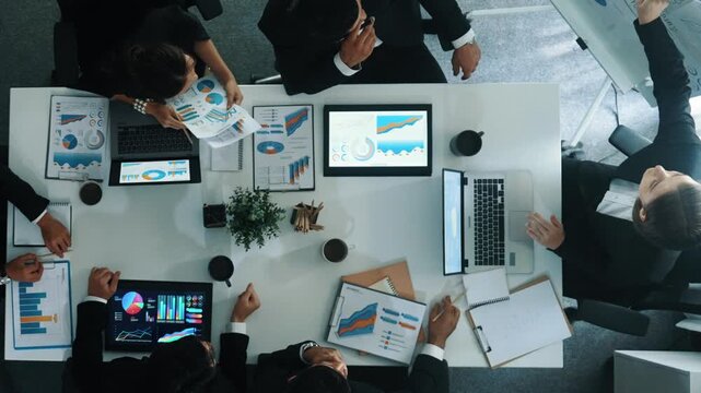 Top down aerial view of smart manager pointing at financial graph on whiteboard while diverse business team explain and plan marketing idea at meeting table with stock market statistic. Directorate.