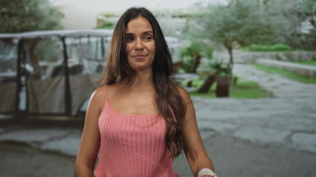 Woman standing in street showing bare shoulders and slight smile, long dark hair, casual pink knit tank, natural skin texture and relaxed posture; confidence.
