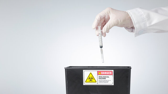 Cautious hand wearing glove drops medical syringe into biohazard container showing responsible clinical waste management disposal technique