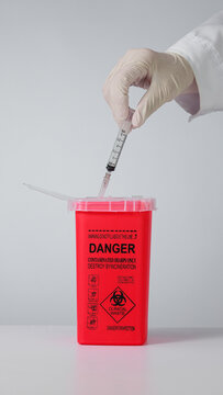 Cautious professional demonstrates safe clinical waste management by dropping used needle syringe into red biohazard sharp disposal container