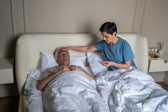 Caring elderly woman checking fever temperature of unwell male partner with thermometer, loving senior wife taking care of ill husband during seasonal cold infection, compassion and family support
