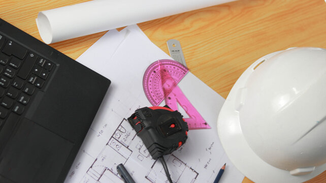 Top view of architect workspace with laptop, hard hat, measuring tape, drafting tools and architectural blueprint on wooden desk. Perfect for engineering, construction and workplace safety concepts.