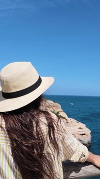 Young woman with long hair wearing a straw hat while enjoying a sunny day on a cliffside, gazing at the beautiful ocean and rocky shoreline from a viewpoint on her summer vacation