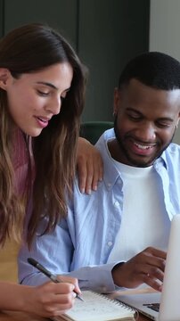 Smiling young multiracial couple working from home, with the man typing on a laptop and the woman taking notes in a notebook while they discuss their project and collaborate as a team