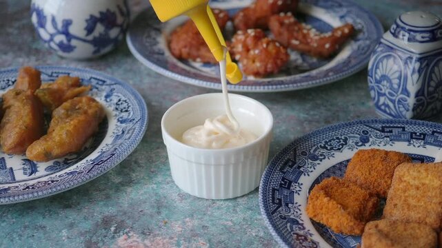 Pouring Creamy Mayonnaise Dip Into Ramekin Beside Fried Appetizers on Blue Patterned Plates