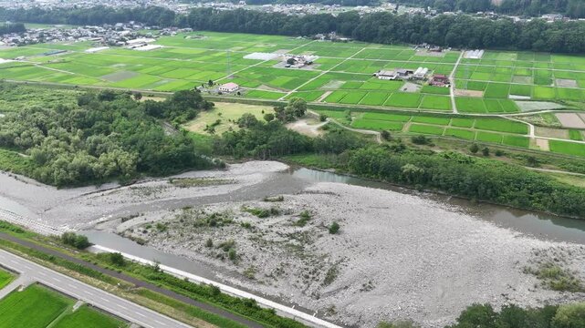 Aerial view shows riverbed between lush rice fields. Gravelly bed runs alongside green paddy rows. Trees frame the landscape, adding depth and texture. Village homes dot the distant horizon