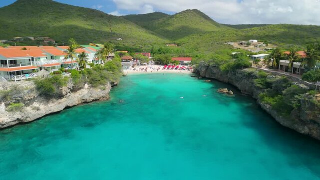 Aerial view of turquoise Playa Lagun cove in Cura&ccedil;ao framed by limestone cliffs, orange roofed villas and palm trees above sandy beach lined with red umbrellas overlooking clear ocean water