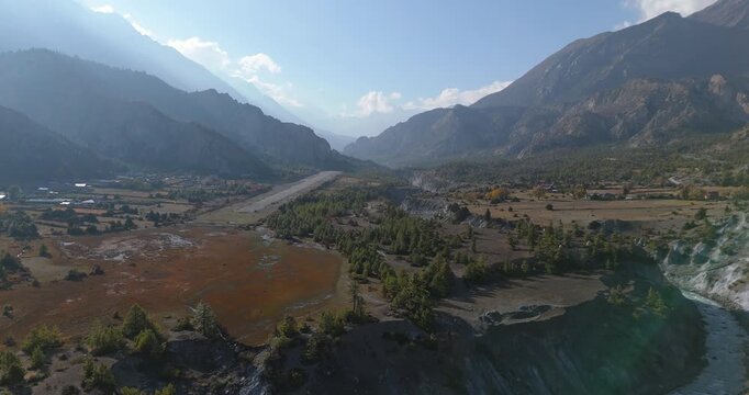 Annapurna Circuit, Manang, Nepal, aerial gliding shot of the winding Marsyangdi River flowing beside Humde Airport airstrip in a wide mountain valley surrounded by Himalayan peaks.