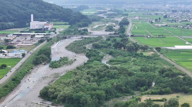 Aerial view of a winding river through green fields. Bare riverbed reveals recent flooding or dry season. Green hills flank a small town with industrial buildings. Bridge crosses the river