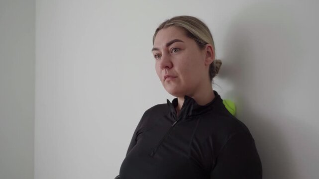 Adult female uses a massage ball against a plain wall to release neck and shoulder trigger points in a simple home setting.