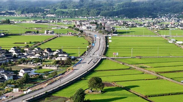 Curving highway winds through lush green rice fields. Village homes dot the landscape beside the road. Vehicles move steadily along the winding route