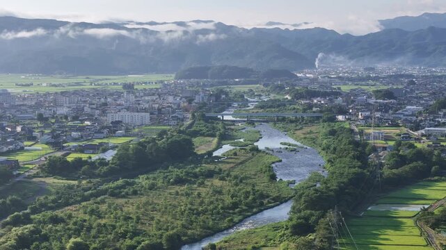 Serene valley wraps around a quiet town. Misty mountains loom behind green fields and rivers. Town lies nestled where water flows gently through fields. Village blends nature with calm human life