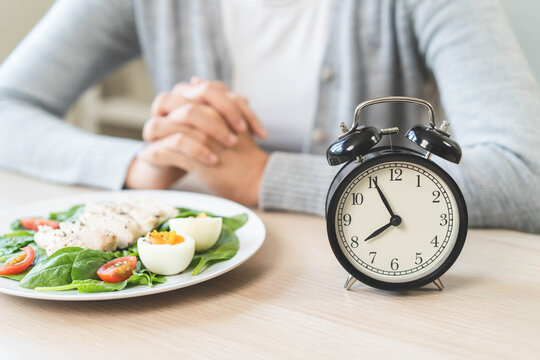 Woman waiting to eat her healthy meal during intermittent fasting concept with alarm clock on wooden table background