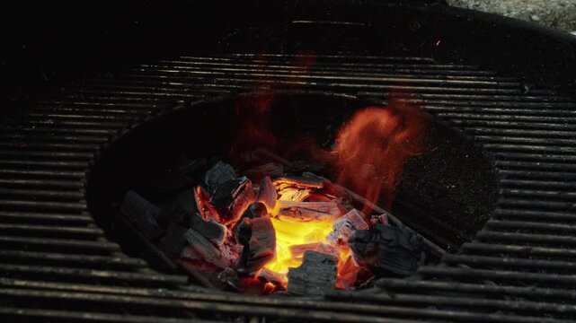 Glowing coals and rising flames burn intensely in the center of a round barbecue grill.