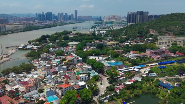 Aerial pullback reveals colorful Coloane Village rooftops along river with distant Macau skyscraper skyline behind