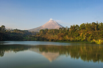Scenic view of Mount Merapi volcano reflected on calm lake water with lush green forest in Central Java, Indonesia. Natural landscape for travel, tourism, conservation, and environmental themes © Hary