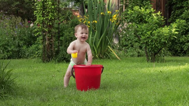 A toddler plays in the backyard. A red bucket is filled with toys and warm water. He takes turns taking out toys and examining them with a smile on his face. Italy