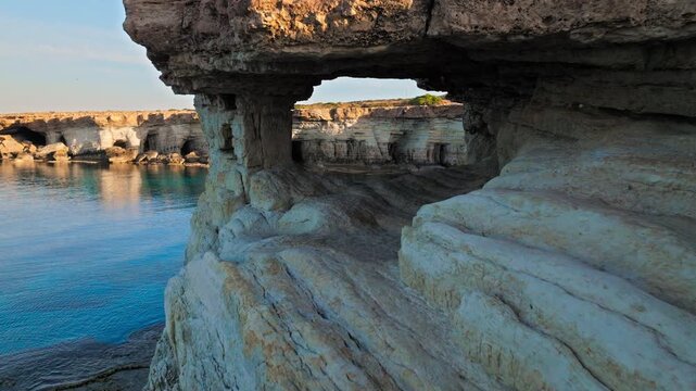 Drone view from inside a sea cave tunnel at Cape Cavo Greco, Cyprus. The arched rock opening frames the blue sea beyond.