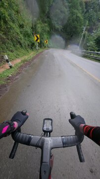 Vertical POV footage of a cyclist riding on a wet mountain road beside a rocky cliff after rain, featuring reflective asphalt, winding curves and scenic tropical landscapes.
