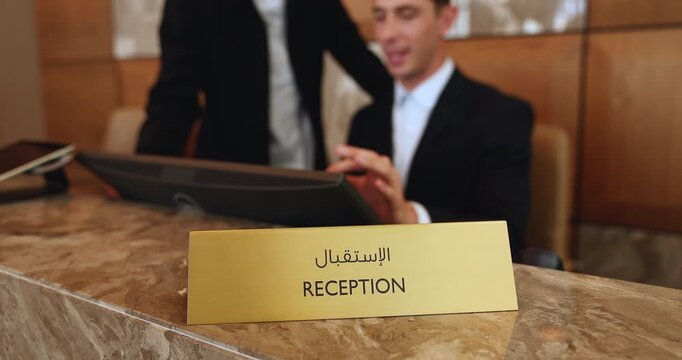 Close up of nameplate with word Reception written in Arabic, placed on hotel reception desk, two receptionists working on guest reservations using pc, representing professional hospitality services