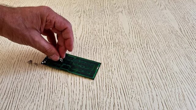A hand places a TV remote control circuit board on a wooden surface, showcasing the green PCB before assembly, repair, testing, or electronic maintenance in a simple workspace