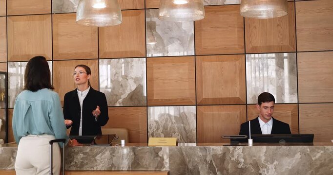 In professional hotel reception staff member assists to woman, guest stands at counter with rolling suitcase makes check-in, get help or information. Tourism, hospitality, efficient customer services