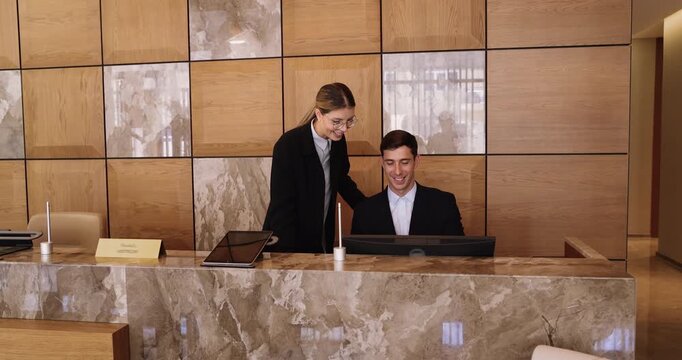 Two receptionists in suits working together at reception desk, managing reservations, entering guest information into system using hotel platform or software. Colleague helps with administrative tasks