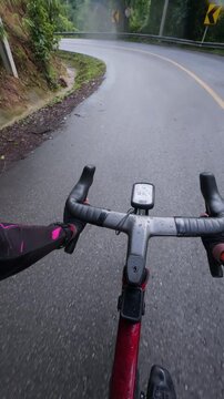 Vertical POV footage of a cyclist descending a wet mountain road after rain, featuring reflective asphalt, smooth curves, guardrails and a scenic tropical forest landscape.