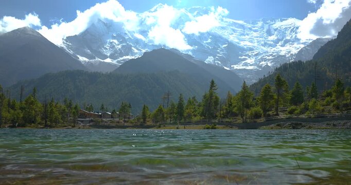 Annapurna Circuit, Manang Nepal, wide camera shot of tranquil green lake water rippling in the foreground with pine forests and snow capped Himalayan peaks in the background.