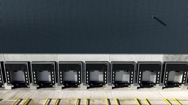 Low-level oblique aerial view tracking past active freight loading docks and roller doors at a modern transport depot.