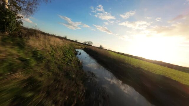 Fast low-altitude FPV drone shot flying along a narrow drainage dyke cutting through vibrant green farming pastures under a dramatic orange sky.