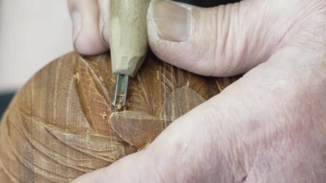 Close up of a carving chisel cutting delicate feather patterns into a wooden surface held in weathered hands